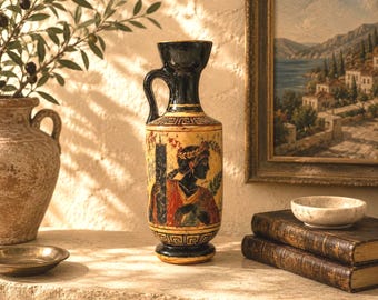 May include: A tall ceramic vase with a black neck and base, featuring a cream-colored body decorated with a black silhouette of a figure wearing a laurel wreath, accented by red and green details and a geometric Greek key pattern around the rim.