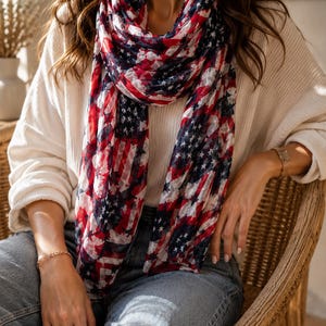 May include: A lightweight scarf featuring a repeating pattern of the American flag with red, white, and blue colors. The scarf is draped around the neck, displaying a print of stars and stripes across the fabric in a soft, semi-sheer material.