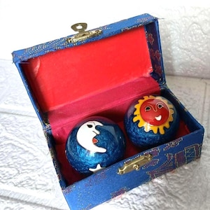 May include: A blue box with a red interior holds two decorative balls. One ball features a white crescent moon, and the other depicts a smiling sun. The box has a gold clasp and is decorated with a blue and gold pattern.
