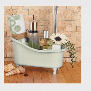Puede incluir: Una pequeña bañera verde claro llena de productos de baño. Contiene una toalla enrollada con lunares verdes, frascos de perfume, una flor blanca y una ramita de vegetación. Conchas marinas y una pequeña piedra están frente a la bañera.