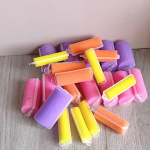 Puede incluir: Un conjunto de rulos de espuma coloridos en tonos rosa, morado, naranja y amarillo. Los rulos son cilíndricos y rectangulares, con una inserción de plástico. El fondo es una superficie de madera clara.