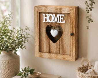 May include: A rectangular wooden wall-mounted key cabinet with a natural wood grain finish. The front features the white text HOME above a heart-shaped cutout containing a small white heart ornament tied with twine. A dark knob is on the right side.