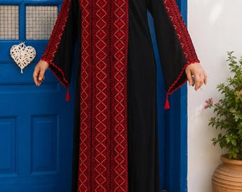 Puede incluir: Un vestido negro hasta el suelo con mangas largas y un panel vertical prominente de bordado geométrico rojo y negro en el centro. Las mangas también presentan bordados rojos a juego, bordes festoneados y pequeñas borlas rojas en los puños.