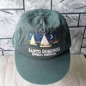Puede incluir: Una gorra de béisbol verde oscuro con una imagen bordada de veleros y una luna creciente. El texto "Santo Domingo Republica Dominicana" está bordado en la parte delantera de la gorra.