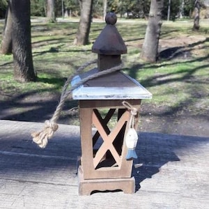 Puede incluir: Linterna de madera con acabado marrón, borde blanco y asa de cuerda. La linterna presenta un diseño en forma de X en los laterales y un pequeño adorno en forma de pez azul y blanco. La linterna es decorativa.