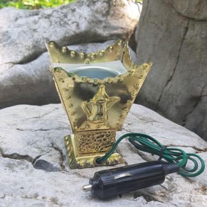 May include: A gold incense burner with a decorative design, featuring a teapot emblem. It has a square base and a pointed top. A black car adapter with a green cord is attached. The burner is on a light-coloured rock. The burner is approximately 15 cm tall.