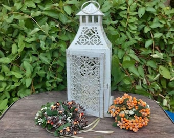 Puede incluir: Una linterna de metal blanco ornamentada, con una parte superior decorativa y paneles laterales de estilo enrejado. Dos pequeños arreglos florales coloridos se encuentran frente a la linterna. El fondo presenta un exuberante follaje verde.