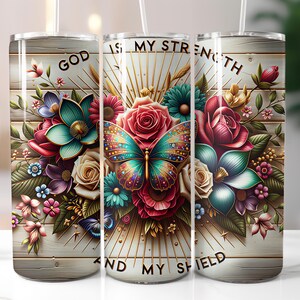 May include: A stainless steel tumbler with a floral design featuring a butterfly, roses, and other flowers. The tumbler has the text "GOD IS MY STRENGTH AND MY SHIELD" printed on it. The background has a wood-like texture.