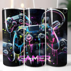 May include: Black tumbler featuring a vibrant design of video game controllers and a headset with colorful paint splatter effects. The word "GAMER" is displayed in a neon font. The tumbler includes a straw.