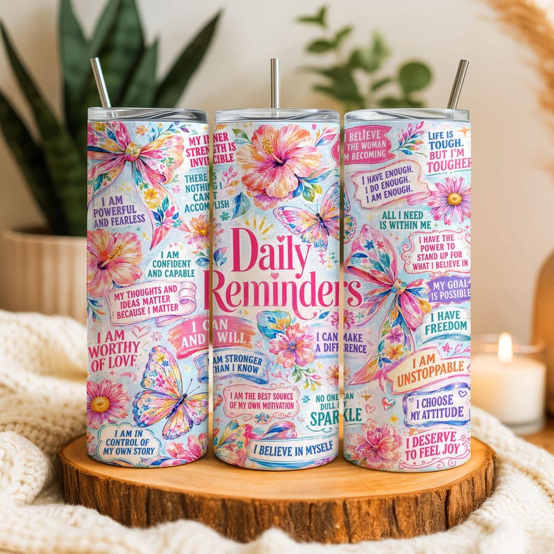 May include: Three stainless steel tumblers with floral and butterfly designs, and the words "Daily Reminders" in pink. Each tumbler has a different inspirational quote. The tumblers are on a wooden surface.