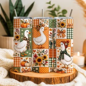 May include: A cylindrical tumbler with a metal straw featuring a patchwork design of white ducks, orange pumpkins, sunflowers, and plaid patterns in shades of green and orange. The surface displays various autumn-themed motifs including bows and hearts.