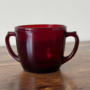 May include: A ruby red glass cup with two curved handles. The cup has a rounded shape and a smooth, glossy finish. The cup is sitting on a wooden surface.