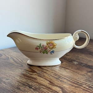 May include: A white gravy boat with a gold rim and floral design. The flowers are yellow, pink, and blue.