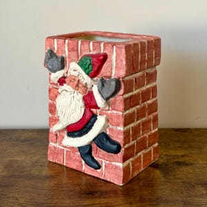 May include: A rectangular, red brick-patterned container with a sculpted Santa Claus climbing on the side. Santa is wearing a red suit, black boots, and a white beard. The container is likely a decorative item for the holiday season.