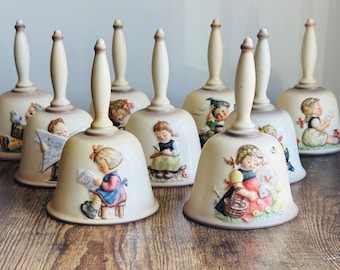 May include: A collection of ceramic bells with cream-colored handles and bell bodies. Each bell features a detailed relief scene of children engaged in various activities, such as reading or playing. The bells are arranged on a wooden surface.