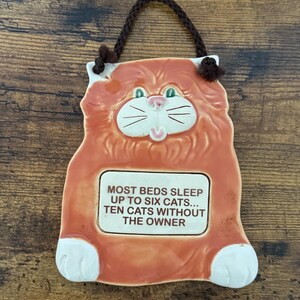 May include: Orange ceramic cat-shaped wall hanging with a brown braided cord for hanging. The cat has a white face, paws, and a rectangular text box that reads, "MOST BEDS SLEEP UP TO SIX CATS... TEN CATS WITHOUT THE OWNER."
