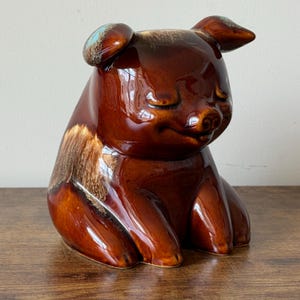 May include: A ceramic pig figurine in a seated position, with a glossy brown glaze. The pig has closed eyes and a small snout, with a touch of blue on the ears. The figurine is a decorative item.