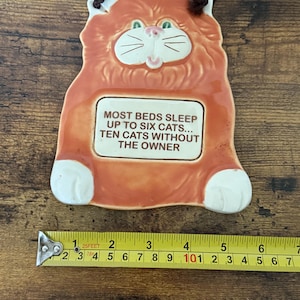 May include: Orange ceramic cat-shaped wall hanging with a brown cord for hanging. The cat has white paws, a white face, and green eyes. Text on the plaque reads: "MOST BEDS SLEEP UP TO SIX CATS... TEN CATS WITHOUT THE OWNER."