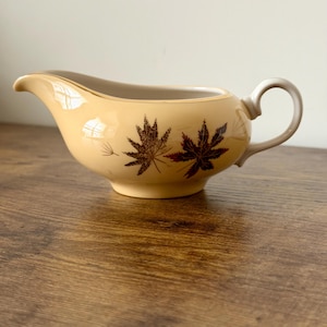 May include: Cream-colored gravy boat with a handle and spout. The boat features a brown leaf design on the side. The interior is a light beige color, and the handle is a light beige color. The gravy boat is made of ceramic.