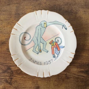 May include: A white ceramic plate with a scalloped edge and gold trim. The plate features a whimsical illustration of two monkeys in blue, red, and yellow. The text "JAMES 1957" is written in black on the plate.