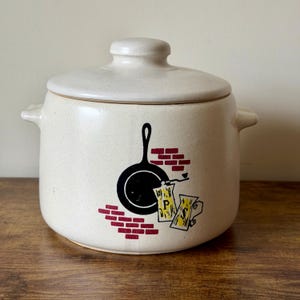 May include: Cream-colored ceramic canister with a lid, featuring a black skillet graphic, red brick accents, and yellow paper scrolls. The canister has side handles and a speckled finish, perfect for kitchen storage.