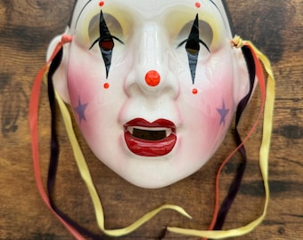 May include: A ceramic clown mask with a glossy white finish, black hair, and painted features including black diamond eye markings, a bright red nose, red lips, and purple stars on the cheeks. The mask includes attached yellow, red, and black ribbons.