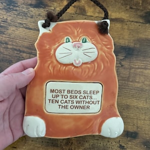 May include: Orange ceramic cat-shaped wall hanging with a brown braided cord for hanging. The cat has white accents, green eyes, and a pink nose. Text on the plaque reads: "MOST BEDS SLEEP UP TO SIX CATS... TEN CATS WITHOUT THE OWNER."
