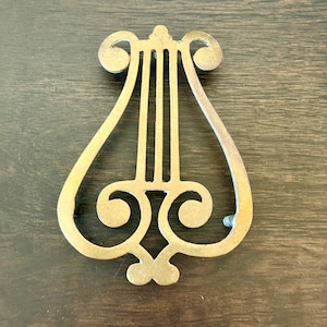 May include: A gold-colored metal lyre-shaped object, featuring a symmetrical design with curved lines and circular accents. The instrument has three vertical bars and is set against a dark wood grain background.
