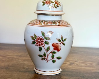 May include: A white ceramic jar with a lid, adorned with floral patterns in red, green, and gold. The jar has a gold rim and a gold finial on the lid. The design is a classic decorative piece.
