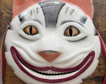 May include: A glossy ceramic cat mask featuring a wide, toothy grin, pointed ears with peach-colored tips, and grey-shaded markings on a white base. The mask has orange almond-shaped eyes and thin ribbons attached to the sides for securing it.