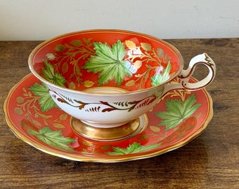 May include: A vintage teacup and saucer set. The cup and saucer are red with green leaf and gold floral designs. The cup has a white interior and handle. The set is on a wooden surface.