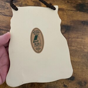 May include: Cream-colored ceramic wall hanging with a brown rope for hanging. The back features a sticker with the text "Smoky Mountain Pottery" and "SMP". The shape is an outline of a mountain range.