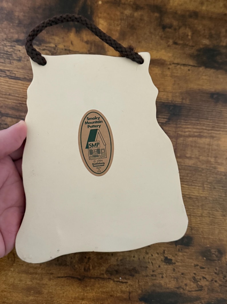 May include: Cream-colored ceramic wall hanging with a brown rope for hanging. The back features a sticker with the text "Smoky Mountain Pottery" and "SMP". The shape is an outline of a mountain range.