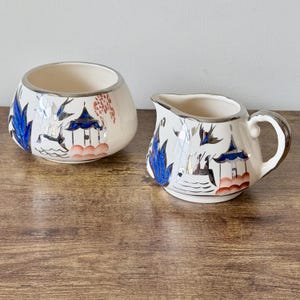 May include: A cream-colored ceramic creamer and sugar bowl set. The set features a silver rim and is decorated with blue, red, and black illustrations of pagodas, birds, and foliage. The sugar bowl is round, and the creamer has a handle and spout.