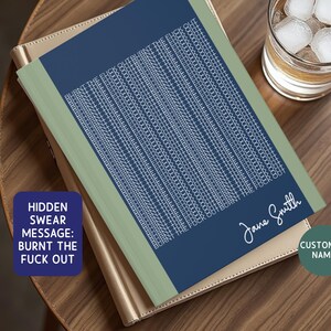 May include: A notebook with a blue cover and a hidden swear message in white text. The notebook has a green border and a gold-colored cover underneath. A glass of whiskey with ice sits next to the notebook on a wooden table.