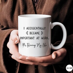 Op de afbeelding: Witte keramische mok met een zwart handvat, met de tekst "I ACCIDENTALLY BECAME IMPORTANT AT WORK. It's Ruining My Life." De mok wordt in de handen van een persoon gehouden. De mok is 325 ml.