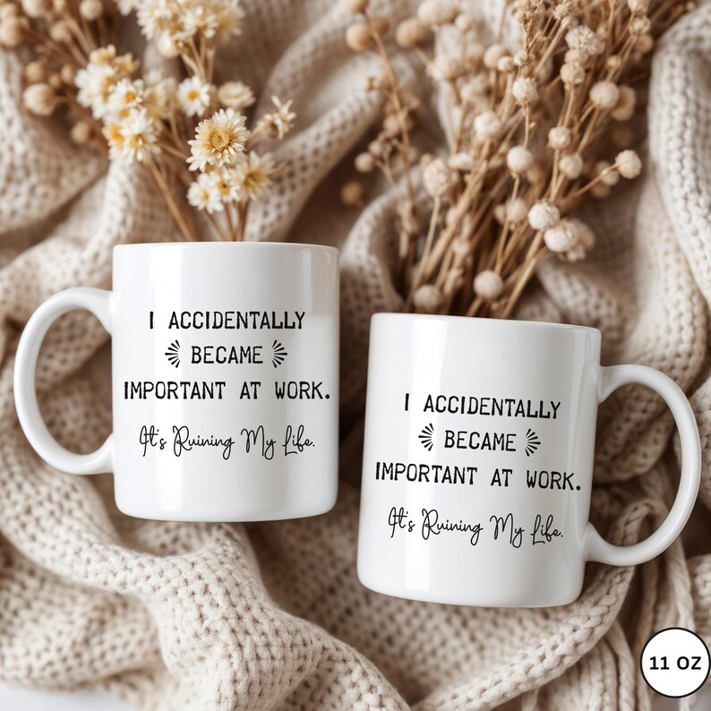 Op de afbeelding: Twee witte keramische mokken met zwarte tekst: "I ACCIDENTALLY BECAME IMPORTANT AT WORK. It's Ruining My Life." De mokken van 312 ml hebben een gebogen handvat. De achtergrond omvat gedroogde bloemen en een beige gebreide deken.