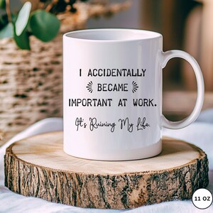 Op de afbeelding: Witte keramische mok met de tekst "I ACCIDENTALLY BECAME IMPORTANT AT WORK. It's Ruining My Life." De mok staat op een houten schijf.