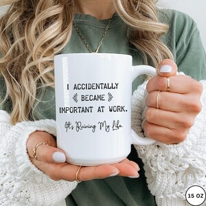 Op de afbeelding: Witte keramische mok met de tekst "I ACCIDENTALLY BECAME IMPORTANT AT WORK. It's Ruining My Life." De mok heeft een zwarte lettertype en een capaciteit van 444 ml. De mok wordt door een persoon vastgehouden.