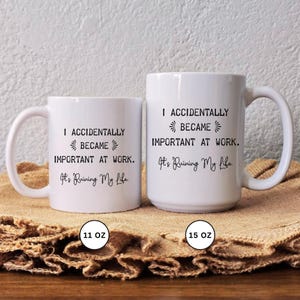 Op de afbeelding: Twee witte keramische mokken met zwarte tekst die luidt: "I ACCIDENTALLY BECAME IMPORTANT AT WORK. It's Ruining My Life." De mokken zijn gelabeld met 11 oz en 15 oz. De mokken staan op een bruin oppervlak.