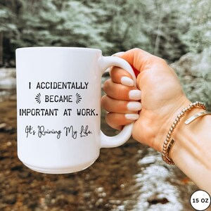 Op de afbeelding: Witte keramische mok met de tekst "I Accidentally Became Important At Work. It's Ruining My Life." De mok heeft een grote handgreep en wordt door een persoon vastgehouden. De mok is 444 ml.