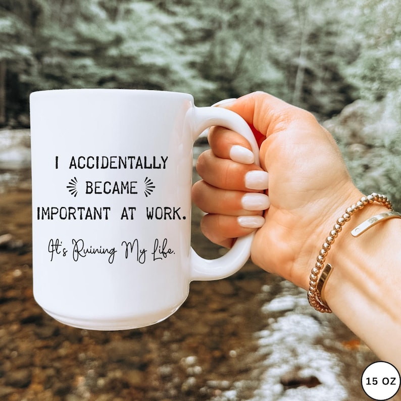 Op de afbeelding: Witte keramische mok met de tekst "I Accidentally Became Important At Work. It's Ruining My Life." De mok heeft een grote handgreep en wordt door een persoon vastgehouden. De mok is 444 ml.