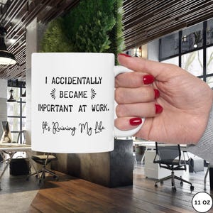 Op de afbeelding: Witte 312g keramische mok met de zwarte tekst "I ACCIDENTALLY BECAME IMPORTANT AT WORK. It's Ruining My Life.". De mok wordt vastgehouden in een hand met rode nagellak. De achtergrond is een kantooromgeving.