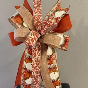 May include: A decorative bow featuring multiple ribbons with a fall theme. The design includes solid burnt orange ribbon, natural tan burlap ribbon, and a cream ribbon printed with orange pumpkins, white gourds, and white botanical leaf patterns.