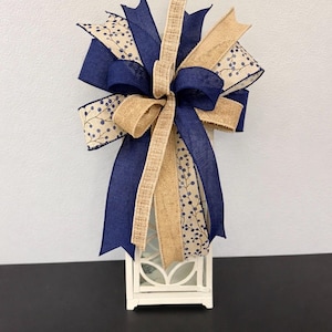 May include: A decorative bow featuring a mix of solid navy blue ribbon, natural tan burlap ribbon, and a cream-colored ribbon with a navy blue berry branch pattern. The bow is arranged in multiple loops and long tails, secured at the center point.