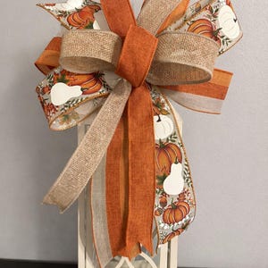 May include: A decorative bow featuring a mix of solid orange fabric, natural tan burlap, and a patterned ribbon displaying orange pumpkins, white gourds, and autumn leaves. The bow is tied with multiple loops and long, trailing ribbon tails.