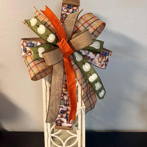 May include: A white metal lantern topped with a decorative multi-layered bow. The bow features ribbons in solid orange, tan burlap, a green pattern with white pumpkins, and a plaid print in orange, yellow, and grey, plus a scarecrow print.
