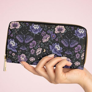 May include: A black wallet with a floral and butterfly pattern in shades of purple and white. The wallet has a gold zipper and is held in a hand.