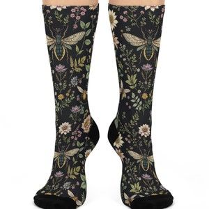 Könnte beinhalten: Ein Paar schwarze Socken mit einem floralen und Insektenmuster. Die Socken zeigen eine Vielzahl von Blumen, Blättern und Insekten, darunter Bienen und Schmetterlinge. Der Druck ist in Grün-, Gelb-, Rosa- und Weißtönen gehalten.