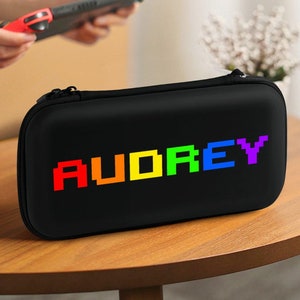 May include: Black carrying case for a Nintendo Switch console with a rainbow pixelated design that says "AUDREY".